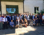 Eine bunt gemischte Gruppe von Menschen, sowohl in Business- als auch in Freizeitkleidung, steht stolz vor einem Gebäude mit einem Schild mit der Aufschrift „Allianz“, das an den akademischen Geist der Hochschule Coburg erinnert. Sie posieren auf Steinpflaster, umgeben von üppigem Grün in der Nähe des Eingangs.