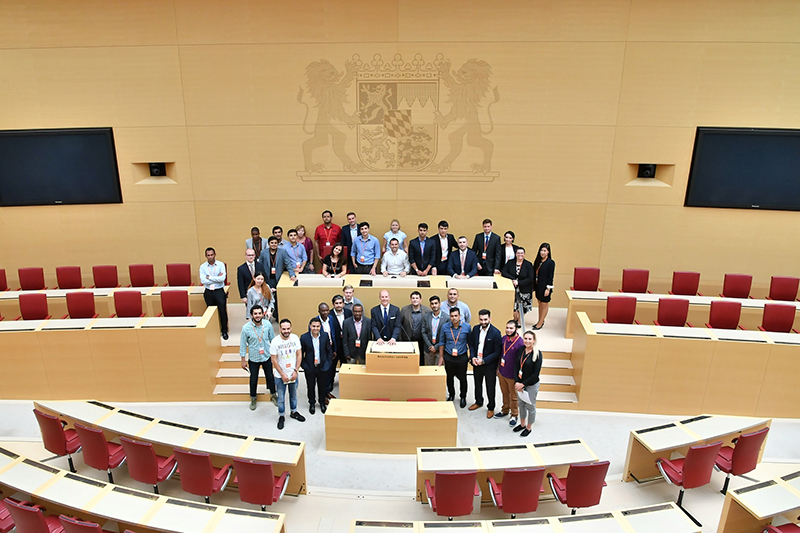 Eine Gruppe von Menschen hat sich in einem hellen Saal mit Holzsitzreihen in einem Parlamentsgebäude versammelt. Der Hintergrund mit einem eingravierten Wappen mit zwei Löwen lässt darauf schließen, dass es sich um eine offizielle Veranstaltung oder ein Treffen handelt, das von der Hochschule Coburg organisiert wird, was auch die professionell gekleideten Teilnehmer vermuten lassen.