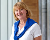 Eine Frau mit kurzen blonden Haaren lächelt. Sie trägt ein weißes Oberteil und einen blauen Pullover über den Schultern, möglicherweise in einer Umgebung der Hochschule Coburg. Sie steht im Innenbereich, wo Holzvertäfelungen und hellgraue Wände die warme Atmosphäre verstärken.