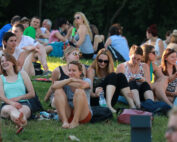 Eine Gruppe von Menschen sitzt bei einer Freiluftveranstaltung der Hochschule Coburg auf einer Wiese und genießt den sonnigen Tag. Einige unterhalten sich, während andere auf ihre Handys schauen oder sich entspannen. Im Hintergrund bilden Bäume eine ruhige Kulisse.