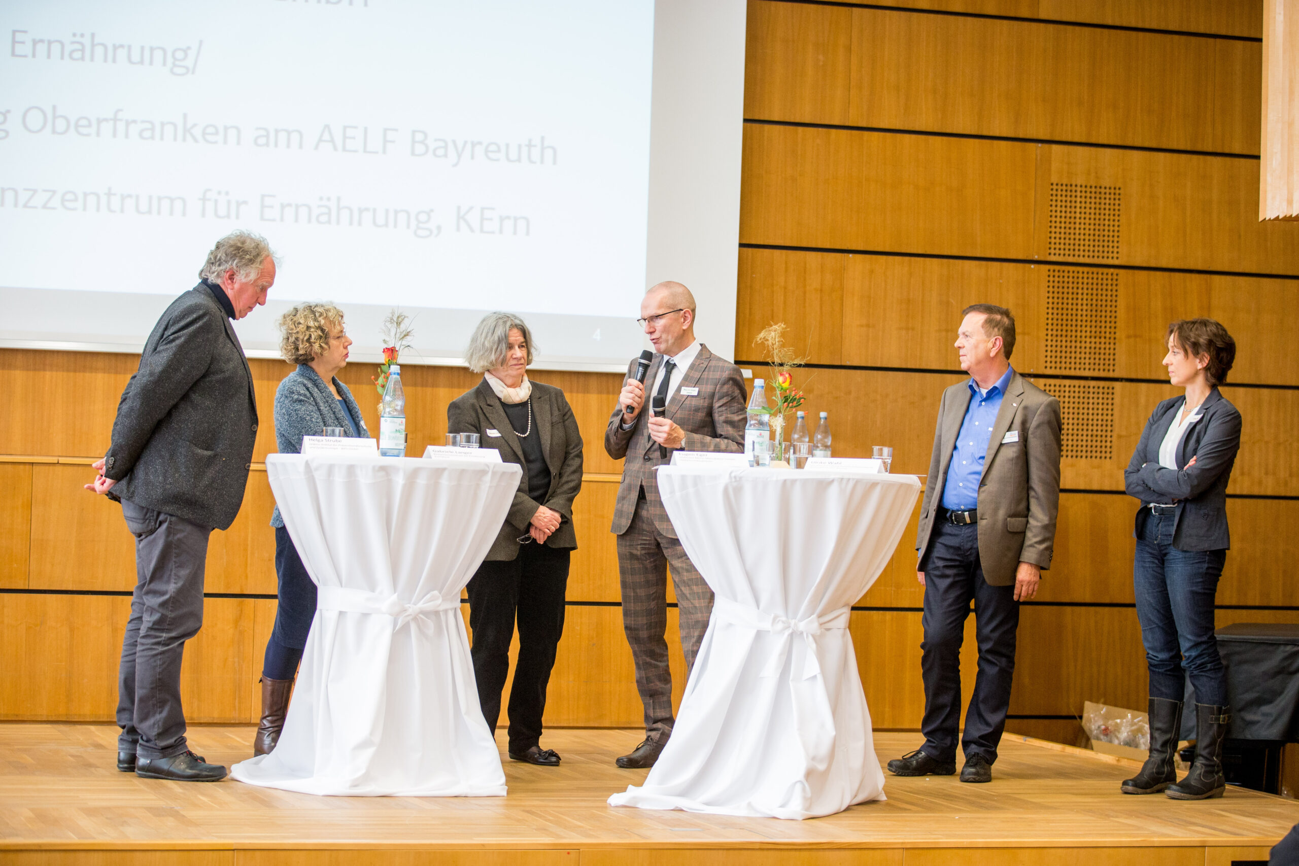 Eine Gruppe von sechs Personen steht auf einer Bühne mit Holzwänden in der Hochschule Coburg und diskutiert mit ihnen. Sie sitzen an zwei kleinen runden Tischen, eine Person hält ein Mikrofon in der Hand.