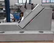 In der Werkstatt der Hochschule Coburg beeindruckt ein Betonprüfstand. Er besteht aus einem schräg geneigten Block, an dem kleinere Bauteile befestigt sind und an dessen Boden Abflüsse zu sehen sind. Im Hintergrund sind Regale und verschiedene Geräte zu sehen, die den Fokus der Einrichtung auf Ingenieursleistung unterstreichen.