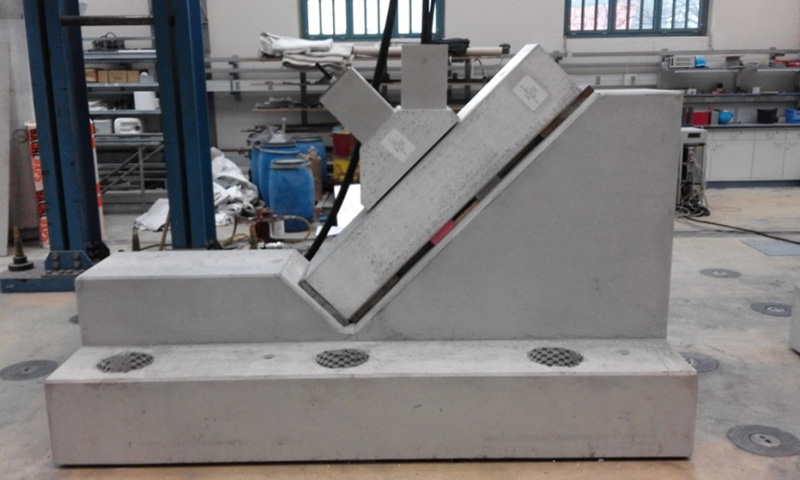 In der Werkstatt der Hochschule Coburg beeindruckt ein Betonprüfstand. Er besteht aus einem schräg geneigten Block, an dem kleinere Bauteile befestigt sind und an dessen Boden Abflüsse zu sehen sind. Im Hintergrund sind Regale und verschiedene Geräte zu sehen, die den Fokus der Einrichtung auf Ingenieursleistung unterstreichen.