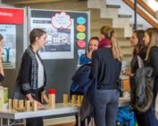 Eine Gruppe von Frauen versammelte sich an einem Tischausstellungsstand der Hochschule Coburg und unterhielt sich zwischen Informationstafeln. Holzklötze und Materialien, die über den Tisch verteilt sind, wecken das Interesse, während nahe gelegene Treppen und teilweise sichtbare Poster auf eine lebendige akademische Umgebung hinweisen.