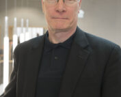 Ein Mann mit Brille, einer dunklen Jacke und einem Hemd mit Kragen steht im Innenbereich der Hochschule Coburg vor einem unscharfen Hintergrund mit vertikalen Lichtern.