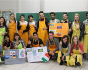 Eine bunt gemischte Gruppe von Studierenden in gelben Schürzen und Freizeitkleidung posiert in einem Hörsaal der Hochschule Coburg. Sie lächeln und strecken den Daumen hoch, während sie vor einer grünen Tafel bunte Plakate über verschiedene Länder, darunter die Türkei und Italien, in die Höhe halten.