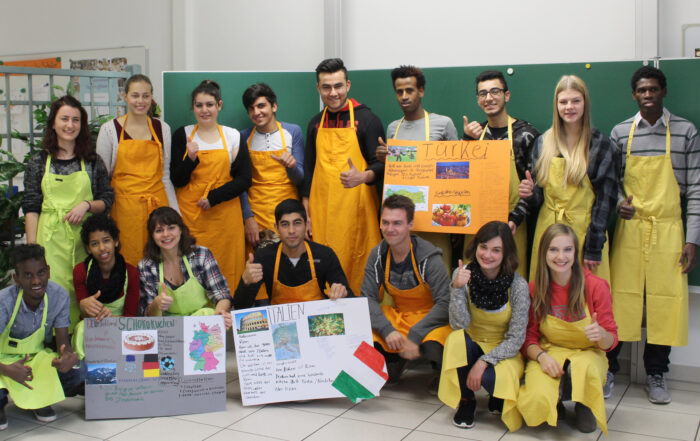 Eine bunt gemischte Gruppe von Studierenden in gelben Schürzen und Freizeitkleidung posiert in einem Hörsaal der Hochschule Coburg. Sie lächeln und strecken den Daumen hoch, während sie vor einer grünen Tafel bunte Plakate über verschiedene Länder, darunter die Türkei und Italien, in die Höhe halten.