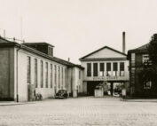 Ein altes Schwarzweißfoto zeigt eine Kopfsteinpflasterstraße mit einem Gebäude mit der Aufschrift „Schlachthof“ in der Mitte. Flankiert von zwei benachbarten Gebäuden, in deren Nähe Fahrräder und Autos geparkt sind, zeigt die Szene Bäume und lässt den historischen Charme in der Nähe der Hochschule Coburg erahnen.