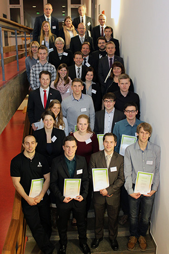 Eine Gruppe von Menschen steht auf einer Treppe der Hochschule Coburg und posiert für ein Foto. Einige halten Zertifikate in den Händen, was darauf schließen lässt, dass es sich um eine Abschluss- oder Preisverleihung handelt. Sie sind formell gekleidet und befinden sich in einem Innenraum mit modernen Designelementen.