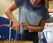In einer geschäftigen Werkstatt bohrt eine Person in blauem Hemd und Jeans geschickt mit einem Elektrowerkzeug Schrauben in ein Holzbrett. Die Kulisse der Hochschule Coburg wird durch Industrieanlagen und Regale im Hintergrund akzentuiert.