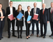 Eine Gruppe von sieben Personen der Hochschule Coburg, alle in formeller Kleidung, steht drinnen und lächelt für ein Foto. Drei Personen halten stolz Ordner in den Händen. Ein Hintergrund mit Logos und dem Text „Nordbayerischer Versicherungstag“ verleiht dem Moment eine offizielle Note.