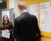 Eine Frau hält eine Tasche und steht neben einem schwarzen Brett, gegenüber einem Mann im Anzug. Das Brett zeigt Plakate mit Texten und Bildern zum Thema Medikamente an der Hochschule Coburg. Im Flur steht ein Schild mit der Aufschrift „Lasst uns mehr erkunden!“.