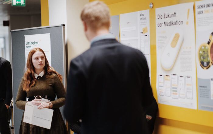 Eine Frau hält eine Tasche und steht neben einem schwarzen Brett, gegenüber einem Mann im Anzug. Das Brett zeigt Plakate mit Texten und Bildern zum Thema Medikamente an der Hochschule Coburg. Im Flur steht ein Schild mit der Aufschrift „Lasst uns mehr erkunden!“.