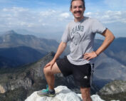 Eine Person steht auf einem Felsen auf einem Berggipfel und trägt stolz ein T-Shirt der Hochschule Coburg und schwarze Shorts. Der Himmel ist klar mit einigen Wolken und gibt den Blick auf die weiten Berge im Hintergrund frei – eine perfekte Mischung aus Natur und Wissenschaft.