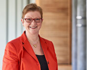 Eine Person mit rotem Blazer und Brille steht lächelnd im Innenbereich der Hochschule Coburg. Im Hintergrund ist ein verschwommenes, modernes Interieur mit hellen Wänden zu sehen.