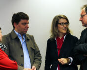 Vier Personen stehen zusammen und unterhalten sich an der Hochschule Coburg. Sie sind in Business-Kleidung gekleidet, eine Frau trägt ein rotes Jackett und einen karierten Schal, die anderen tragen dunklere Anzüge. Der Raum ist vor einem schlichten Hintergrund.