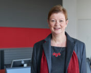 Eine Person mit kurzen Haaren, einem grauen Blazer und einer roten Halskette steht lächelnd in einem Büro der Hochschule Coburg. Im Hintergrund sind rot-graue Wände und Computerstationen zu sehen, die den akademischen Aspekt mit modernem Design perfekt verbinden.