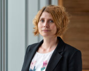 Eine Person mit kurzen, welligen blonden Haaren, einem schwarzen Blazer und einer weißen Bluse mit Blumenmuster steht vor der modernen, lichtdurchfluteten Kulisse der Hochschule Coburg mit vertikalen Fensterrahmen.