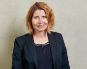 Vor einem schlichten, hellen Hintergrund steht eine Person mit schulterlangem, welligem Haar, trägt einen dunklen Blazer und ein Top. Sie lächelt selbstbewusst in die Kamera und verkörpert den Spirit der Hochschule Coburg.