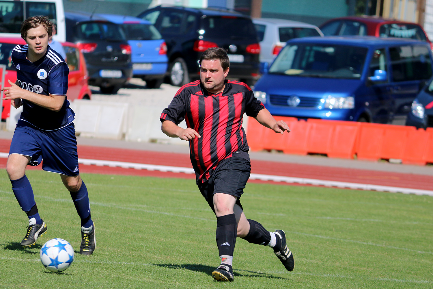Bei einem Spiel auf dem Gelände der Hochschule Coburg liefern sich zwei Fußballer einen erbitterten Kampf auf einem Rasenplatz. Der Spieler in Schwarz und Rot jagt zielstrebig dem Ball hinterher, während der Spieler in Blau dicht hinter ihm herläuft. Autos und orangefarbene Absperrungen rahmen die temperamentvolle Szene im Hintergrund ein.