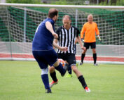 Auf einem Rasenplatz der Hochschule Coburg kämpfen zwei Fußballspieler um den Ball. Einer trägt ein blaues Trikot, der andere ein schwarz-weiß gestreiftes Trikot. Im Hintergrund sind ein Torwart in einem orangefarbenen Trikot und ein weiterer Spieler in der Nähe des Tores zu sehen.
