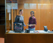 Zwei Frauen stehen lächelnd in einer Werkstatt der Hochschule Coburg hinter einer Glasscheibe. Eine hält ein Werkzeug in der Hand, während ein 3D-Drucker, ein Stapel Kabel und andere Geräte die hölzerne Werkbank vor ihnen überladen. Regale und Schränke säumen ordentlich die holzgetäfelten Wände.
