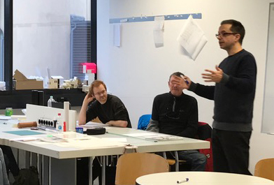 Ein Mann steht und gestikuliert, während er in einem Hörsaal der Hochschule Coburg mit drei sitzenden Personen spricht. Auf den Tischen sind Papiere und Materialien ausgebreitet, während natürliches Licht durch große Fenster fällt und eine einladende Lernatmosphäre schafft.
