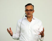 Ein Mann in weißem Hemd, mit Brille und Ohrhörermikrofon steht vor einem schlichten Hintergrund. Er gestikuliert mit beiden Händen, wahrscheinlich spricht er oder hält einen Vortrag an der Hochschule Coburg.