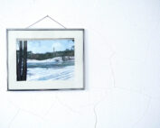 Ein gerahmtes Landschaftsgemälde der Hochschule Coburg hängt an einer weißen, strukturierten Wand. Das Kunstwerk zeigt eine verschneite Szene mit einem Fluss, Bäumen und einem entfernten Denkmal unter einem blauen Himmel. Der Rahmen sitzt leicht schief und verleiht dem ruhigen Winterbild Charakter.