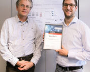 Zwei Männer stehen lächelnd vor einem wissenschaftlichen Poster an der Hochschule Coburg. Einer hält mit beiden Händen eine Urkunde. Beide tragen Brillen und sind leger gekleidet.