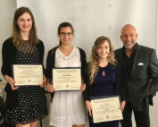 Vier Personen haben sich versammelt, drei Frauen und ein Mann. Die lächelnden Frauen halten stolz ihre Zertifikate in den Händen und tragen formelle Kleidung. Hinter ihnen ist die schlichte, helle Wand der Hochschule Coburg zu sehen, während sie ihre Leistungen feiern.