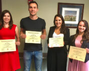 Vier Personen stehen in einem Gebäude der Hochschule Coburg, lächeln und halten Zertifikate in den Händen. Sie tragen halbformelle Kleidung, im Hintergrund hängen gerahmte Kunstwerke an der Wand.