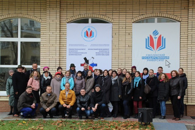 Eine Gruppe von Menschen posiert vor einem Backsteingebäude, das mit großen Bannern geschmückt ist, auf denen der Name „Hochschule Coburg“ und ein Universitätslogo in russischer Sprache zu sehen sind. Warm angezogen, was auf einen kalten Tag schließen lässt, knien oder sitzen einige Mitglieder in der ersten Reihe.