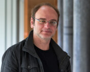 Eine Person mit Brille, schwarzem Hemd und Jackett steht mit einem leichten Lächeln im Flur der Hochschule Coburg, umgeben von weißen Wänden und Säulen. Die kurzen Haare der Person ergänzen das akademische Ambiente perfekt.