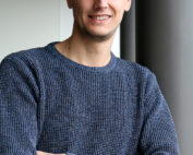 Eine Person mit kurzen dunklen Haaren steht lächelnd mit verschränkten Armen da und trägt einen blauen Strickpullover. Sie lehnt an einer dunklen Säule im Inneren der Hochschule Coburg, im Hintergrund herrscht helles Treiben im Inneren.