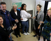 Eine Gruppe von sechs Personen steht in einem Raum der Hochschule Coburg, hält Gläser mit Wasser in den Händen und lächelt. Sie haben sich um einen Wasserspender versammelt, im Hintergrund sind Plakate und ein Getränkeautomat zu sehen.