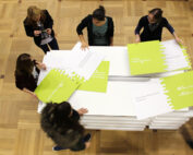 Eine Gruppe von Menschen, die den Geist der Hochschule Coburg verkörpern, steht um einen Tisch und ordnet große Puzzleteile. Die grünen rechteckigen Teile mit weißem Text liegen auf einem Holzboden und fördern eine Atmosphäre, die sowohl kollaborativ als auch konzentriert ist.