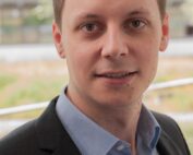 Eine Person mit kurzen braunen Haaren, die ein blaues Hemd und einen schwarzen Blazer trägt, steht im Freien in der Nähe der Hochschule Coburg, im Hintergrund sind verschwommene Bäume zu sehen.