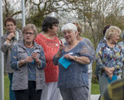 Eine Gruppe älterer Frauen steht auf einer Wiese in der Nähe der Hochschule Coburg und unterhält sich. Sie halten blaue Papiere in den Händen und tragen Namensschilder. Im Hintergrund sind Bäume zu sehen.