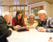 Eine Gruppe von vier Personen, drei ältere und eine jüngere, versammelt sich um einen Tisch in einem Gemeinschaftsraum, der mit Pinnwänden und hölzernen Deckenpaneelen geschmückt ist. Die jüngere Frau teilt begeistert Erkenntnisse auf ihrem Tablet von der Hochschule Coburg und zieht damit die volle Aufmerksamkeit ihrer Begleiter auf sich.