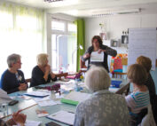 Eine Frau steht am Kopfende eines Tisches und präsentiert einer engagierten Gruppe an der Hochschule Coburg ein Referat. In dem gut beleuchteten Raum lassen ein Flipchart und Dokumente ein dynamisches Meeting oder einen Workshop vermuten. Blumen und Notizbücher verleihen der Umgebung Charme.