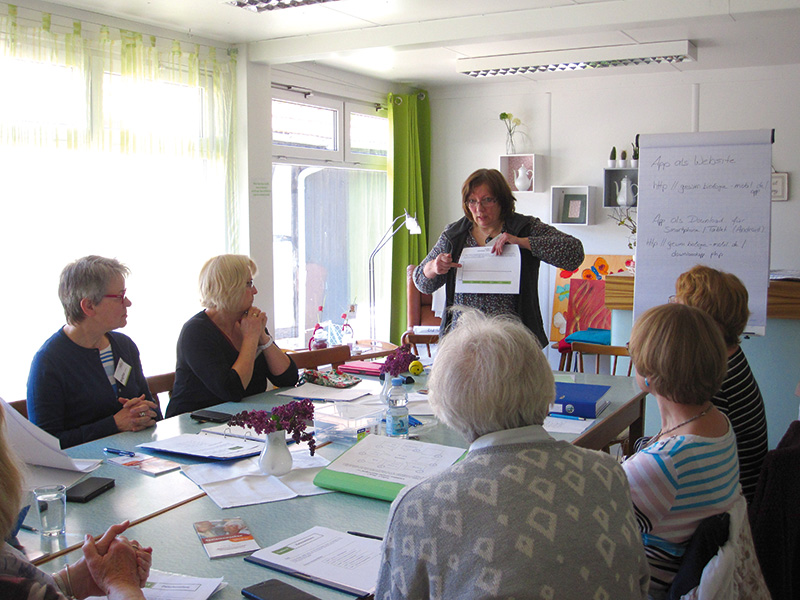 Eine Frau steht am Kopfende eines Tisches und präsentiert einer engagierten Gruppe an der Hochschule Coburg ein Referat. In dem gut beleuchteten Raum lassen ein Flipchart und Dokumente ein dynamisches Meeting oder einen Workshop vermuten. Blumen und Notizbücher verleihen der Umgebung Charme.