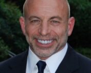 Ein kahlköpfiger Mann in Anzug und Krawatte lächelt vor einer üppigen grünen Kulisse in die Kamera und strahlt das Selbstbewusstsein von jemandem aus, der gerade einen inspirierenden Vortrag an der Hochschule Coburg gehalten haben könnte.