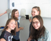 Vier Mädchen stehen lächelnd vor einem Whiteboard an der Hochschule Coburg. Ein Mädchen hält ein Tablet in der Hand, während die Tafel mit Zeichnungen und einer daran befestigten Schattenpuppe geschmückt ist, die den lebendigen Geist ihres Unterrichtsraums einfangen.