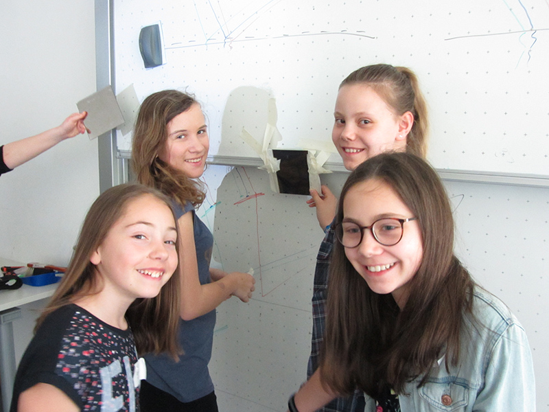 Vier Mädchen stehen lächelnd vor einem Whiteboard an der Hochschule Coburg. Ein Mädchen hält ein Tablet in der Hand, während die Tafel mit Zeichnungen und einer daran befestigten Schattenpuppe geschmückt ist, die den lebendigen Geist ihres Unterrichtsraums einfangen.