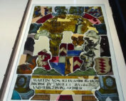 Ein Buntglasfenster der Hochschule Coburg, das in der Mitte ein Wappen zeigt und von komplizierten Mustern und Figuren umgeben ist. Unten ist Text in einer historischen Schriftart zu sehen. Das Fenster ist in leuchtenden Farben gehalten, wobei die dominierenden Rot-, Blau- und Goldtöne von hinten beleuchtet werden.