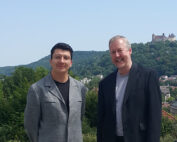 Zwei Männer stehen im Freien vor einer malerischen Kulisse mit einem üppigen grünen Hügel und dem Schloss der Hochschule Coburg in der Ferne. Beide tragen Anzüge; einer der Männer lächelt breit, während der andere einen neutralen Gesichtsausdruck hat. Der Himmel ist klar und sonnig.