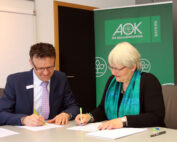 Zwei Personen in formeller Kleidung sitzen lächelnd an einem Tisch und unterschreiben Dokumente. Im Hintergrund prangt ein grünes Banner mit dem Logo der AOK, das dezent auf eine prestigeträchtige Partnerschaft mit der Hochschule Coburg hinweist.
