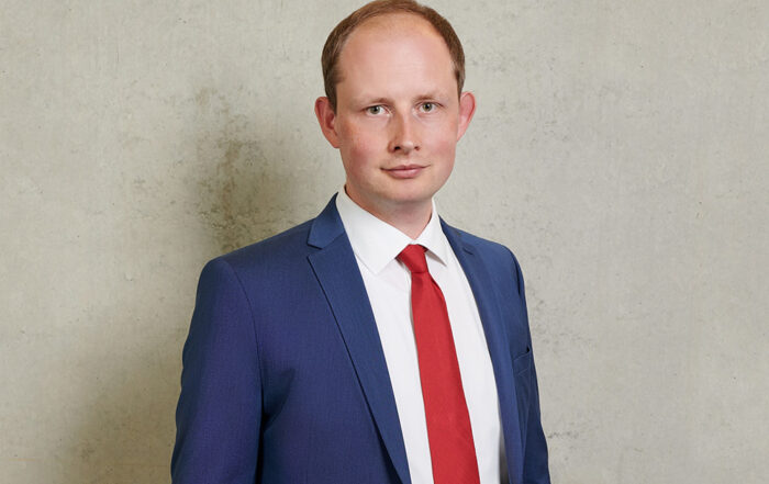 Eine Person in blauem Anzug und roter Krawatte steht vor einem grauen Betonhintergrund und verkörpert die Professionalität, die mit der Hochschule Coburg verbunden wird. Sie blickt mit neutralem Gesichtsausdruck in die Kamera.