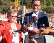 Zwei Personen in formeller Kleidung benutzen während einer Freiluftveranstaltung an der Hochschule Coburg Schlägel. Die Person links trägt eine rote Jacke und die Person rechts einen blauen Anzug und eine Krawatte. Andere Personen mit ähnlichen Schlägeln nehmen an dieser zeremoniellen Aktivität teil.